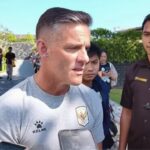 Man in a light gray Kelme shirt being interviewed by reporters outdoors, smartphones visible in foreground.