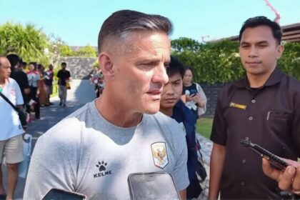 Man in a light gray Kelme shirt being interviewed by reporters outdoors, smartphones visible in foreground.