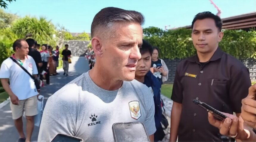 Man in a light gray Kelme shirt being interviewed by reporters outdoors, smartphones visible in foreground.
