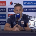Man in a dark blue jersey speaks into a microphone at a press conference, with a soccer ball on the table and sponsor banners in the background.