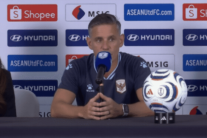 Man in a dark blue jersey speaks into a microphone at a press conference, with a soccer ball on the table and sponsor banners in the background.