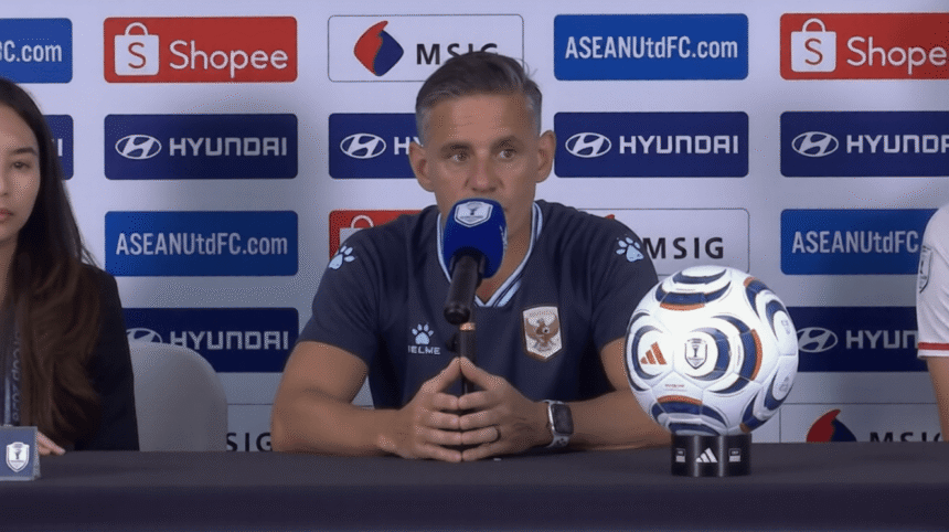 Man in a dark blue jersey speaks into a microphone at a press conference, with a soccer ball on the table and sponsor banners in the background.