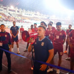 Coach with a red megaphone addresses a line of maroon-clad football players on a track inside a crowded stadium gathered for a ceremony or briefing.