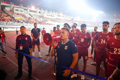 Coach with a red megaphone addresses a line of maroon-clad football players on a track inside a crowded stadium gathered for a ceremony or briefing.