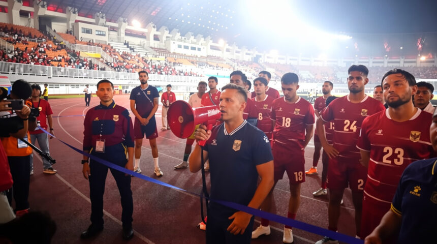 Coach with a red megaphone addresses a line of maroon-clad football players on a track inside a crowded stadium gathered for a ceremony or briefing.
