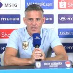 Man in light-blue polo speaks into a blue microphone at a press conference; nameplate reads John Herdman, Head Coach, with a soccer ball on the table and sponsor logos in the background.