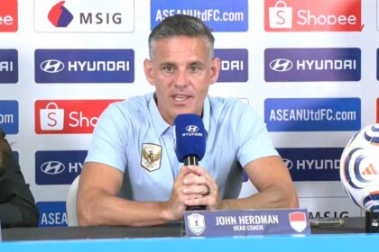 Man in light-blue polo speaks into a blue microphone at a press conference; nameplate reads John Herdman, Head Coach, with a soccer ball on the table and sponsor logos in the background.
