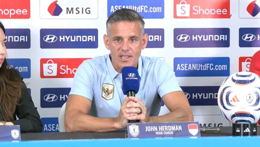 Man in light-blue polo speaks into a blue microphone at a press conference; nameplate reads John Herdman, Head Coach, with a soccer ball on the table and sponsor logos in the background.