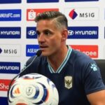 Male soccer manager at a press conference, speaking into a microphone with a branded soccer ball in front, backdrop featuring sponsors like Hyundai and Shopee