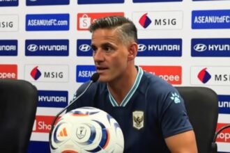 Male soccer manager at a press conference, speaking into a microphone with a branded soccer ball in front, backdrop featuring sponsors like Hyundai and Shopee