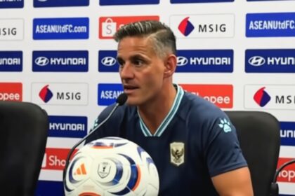 Male soccer manager at a press conference, speaking into a microphone with a branded soccer ball in front, backdrop featuring sponsors like Hyundai and Shopee