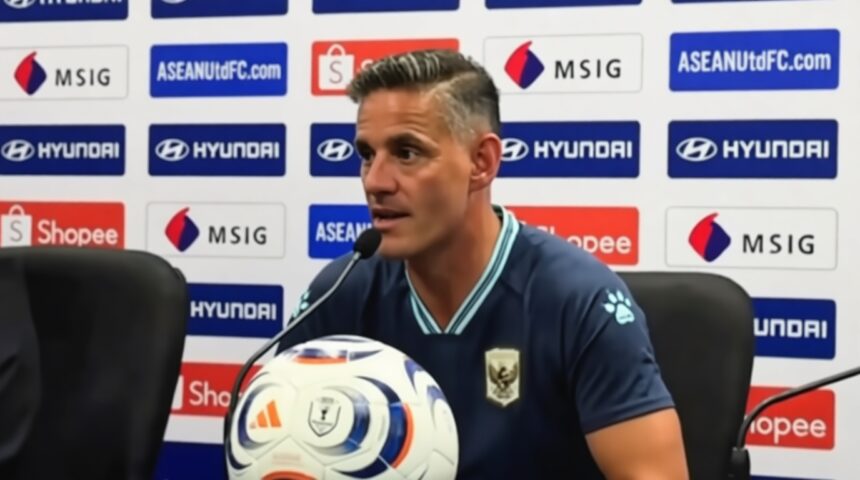 Male soccer manager at a press conference, speaking into a microphone with a branded soccer ball in front, backdrop featuring sponsors like Hyundai and Shopee
