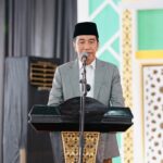 Man in a gray suit and black cap delivering a speech at a lectern on a stage with a microphone.