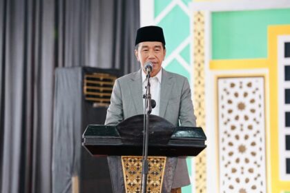 Man in a gray suit and black cap delivering a speech at a lectern on a stage with a microphone.