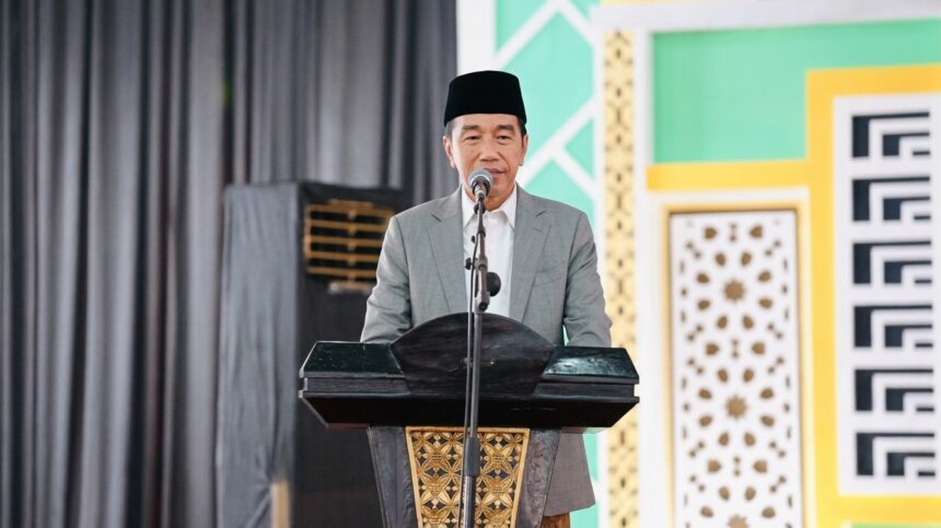 Man in a gray suit and black cap delivering a speech at a lectern on a stage with a microphone.
