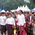 A group of officials and attendees in traditional Indonesian clothing wave as they walk past a crowd under white festival tents.