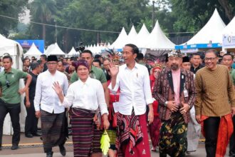 A group of officials and attendees in traditional Indonesian clothing wave as they walk past a crowd under white festival tents.