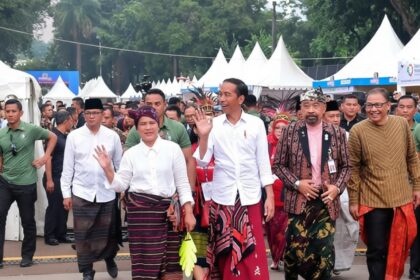 A group of officials and attendees in traditional Indonesian clothing wave as they walk past a crowd under white festival tents.