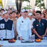 Group of men in white and dark shirts standing behind a table at a crowded outdoor event, water bottles and fruit on the table ahead of them.