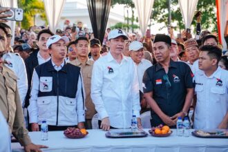 Group of men in white and dark shirts standing behind a table at a crowded outdoor event, water bottles and fruit on the table ahead of them.