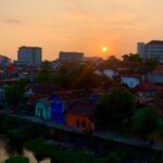 Sunset over a river town with colorful houses and trees, distant modern buildings on the horizon.