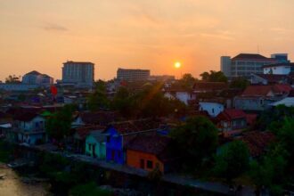 Sunset over a river town with colorful houses and trees, distant modern buildings on the horizon.
