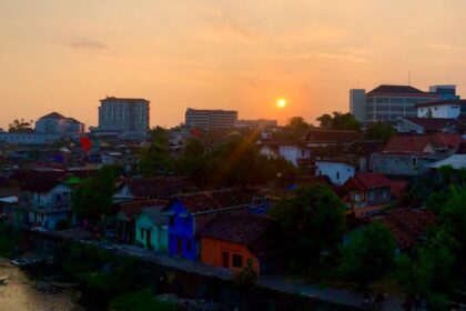 Sunset over a river town with colorful houses and trees, distant modern buildings on the horizon.