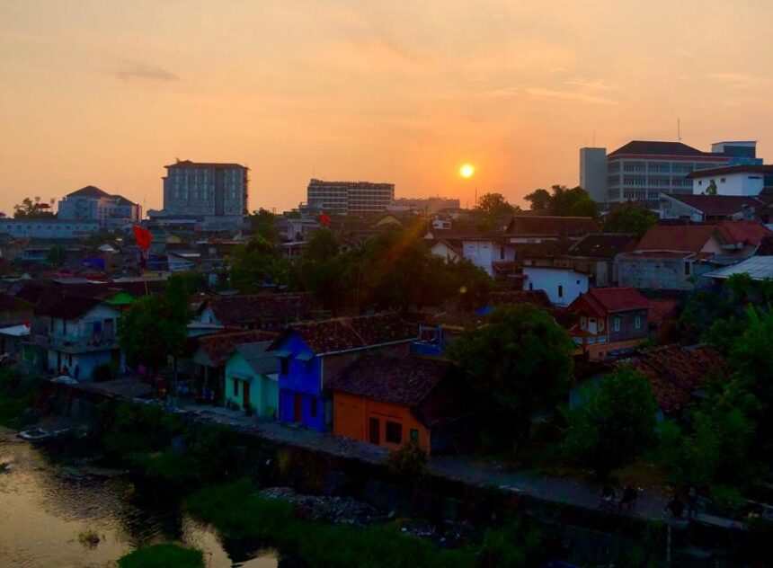 Sunset over a river town with colorful houses and trees, distant modern buildings on the horizon.