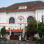 White two-story building with orange trim, arched windows, and a central clock; street, trees, flags, and pedestrians in front.