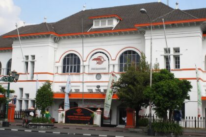 White two-story building with orange trim, arched windows, and a central clock; street, trees, flags, and pedestrians in front.