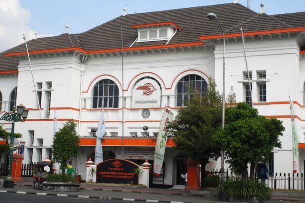 White two-story building with orange trim, arched windows, and a central clock; street, trees, flags, and pedestrians in front.