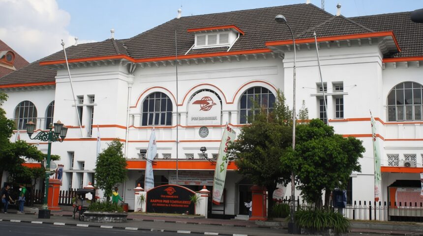 White two-story building with orange trim, arched windows, and a central clock; street, trees, flags, and pedestrians in front.