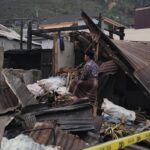 Man in traditional clothing stands amid collapsed debris and wrecked buildings, surveying the destruction behind yellow caution tape.