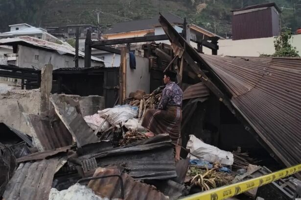 Man in traditional clothing stands amid collapsed debris and wrecked buildings, surveying the destruction behind yellow caution tape.