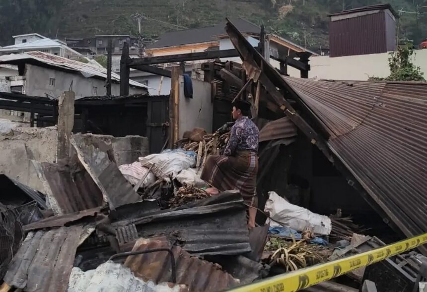 Man in traditional clothing stands amid collapsed debris and wrecked buildings, surveying the destruction behind yellow caution tape.