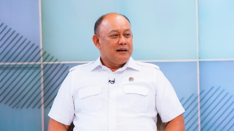 Man in a white short-sleeve shirt with a lavalier mic, seated and talking in a studio with blue panels behind him and diagonal lines.