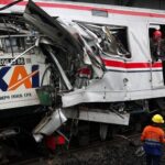 Front of a damaged passenger train with crushed metal; workers in helmets assess the wreckage on the tracks.