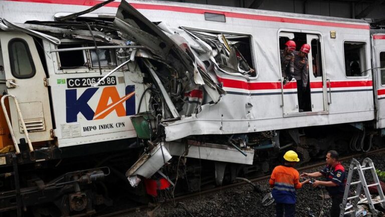 Front of a damaged passenger train with crushed metal; workers in helmets assess the wreckage on the tracks.