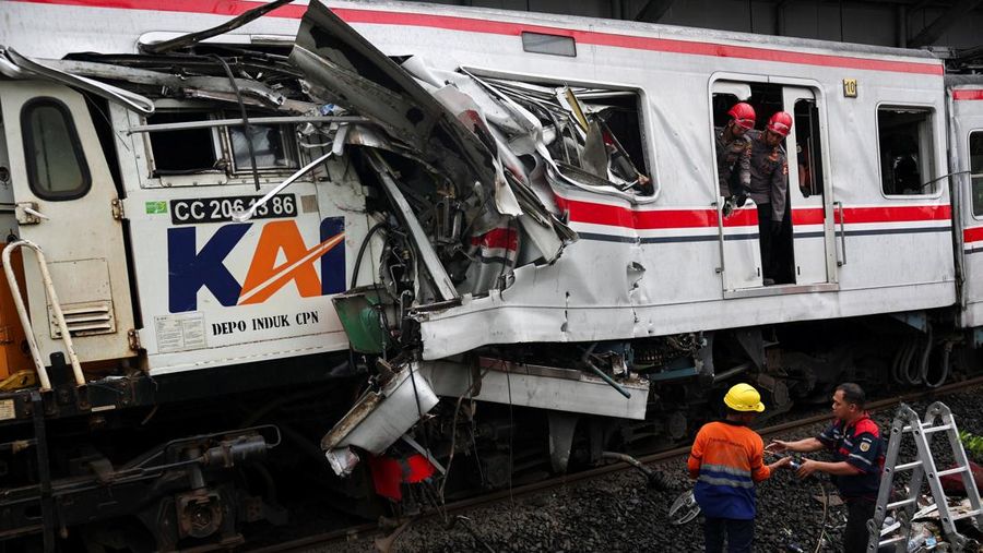 Front of a damaged passenger train with crushed metal; workers in helmets assess the wreckage on the tracks.