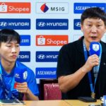 Two men sit at a press conference table with sponsor banners behind; left is a young player in a blue polo, right is a coach speaking into a microphone.