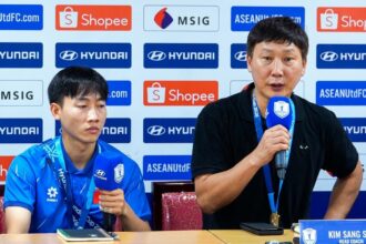 Two men sit at a press conference table with sponsor banners behind; left is a young player in a blue polo, right is a coach speaking into a microphone.