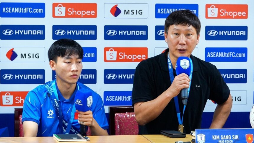 Two men sit at a press conference table with sponsor banners behind; left is a young player in a blue polo, right is a coach speaking into a microphone.