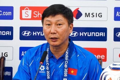 Man in a blue polo speaking into a microphone at a press conference, backdrop with sponsor logos and a Vietnamese flag on his chest.