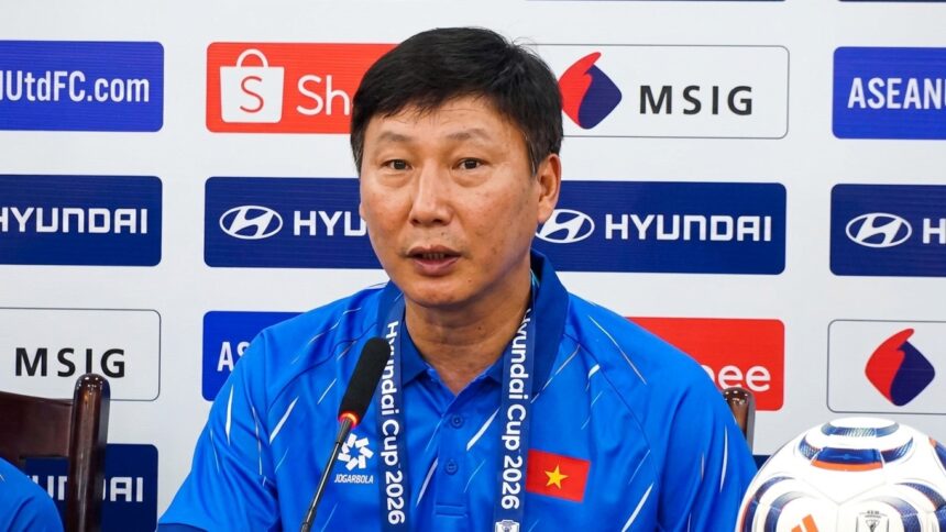 Man in a blue polo speaking into a microphone at a press conference, backdrop with sponsor logos and a Vietnamese flag on his chest.