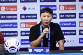 Press conference scene: man in black shirt speaks into a microphone at a table, backdrop full of sponsor logos; nameplate reads Kim Sang Sik.