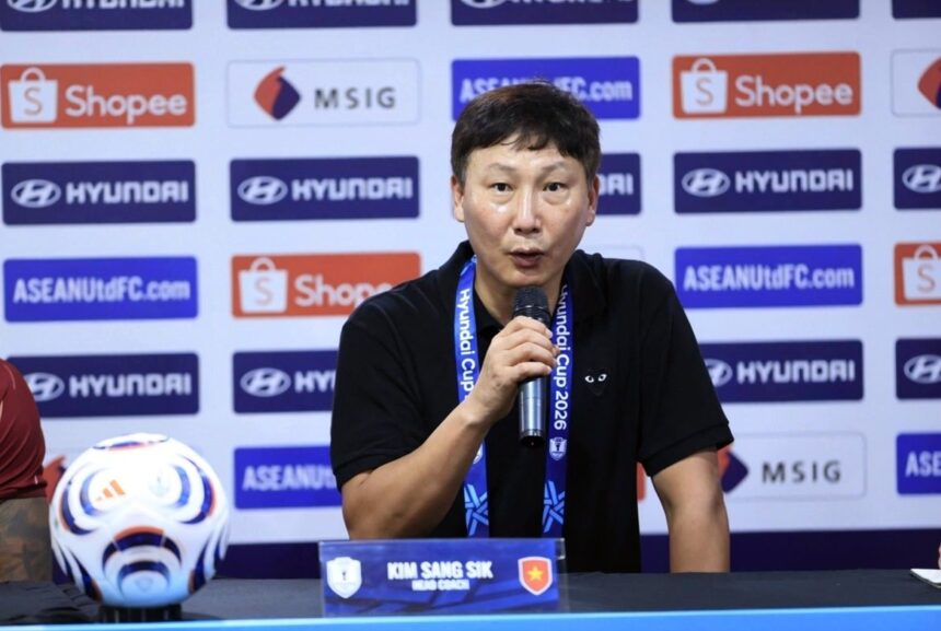 Press conference scene: man in black shirt speaks into a microphone at a table, backdrop full of sponsor logos; nameplate reads Kim Sang Sik.