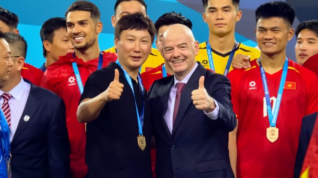 Group of medal-winning athletes posing for a photo, two men in front giving thumbs up in suits and black shirt with gold medals on lanyards