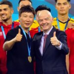 Group of medal-winning athletes posing for a photo, two men in front giving thumbs up in suits and black shirt with gold medals on lanyards