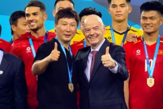 Group of medal-winning athletes posing for a photo, two men in front giving thumbs up in suits and black shirt with gold medals on lanyards