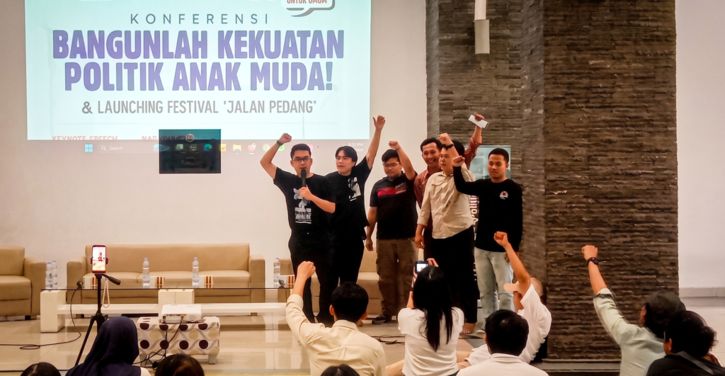 Konferensi Politik Anak Muda FIXX - Mabur.co Group of young men on a stage raising their fists in celebration during a conference, with a large banner behind them reading a political youth theme in Indonesian.